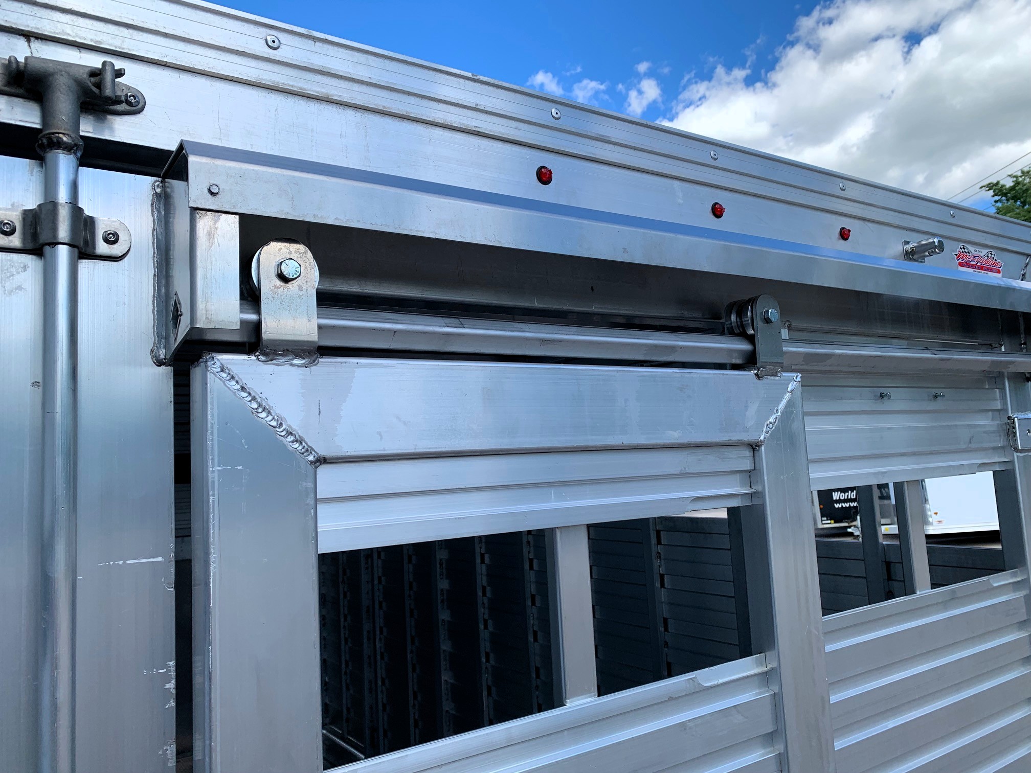 image8 15 - McFarlane Trailer Sales Close-up of a silver aluminum trailer side with a hinge, latch, and small red marker lights under a blue sky.