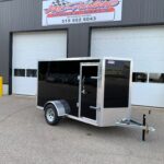 Black enclosed utility trailer with diamond-plate front parked in front of a grey industrial building with Roll-Up garage doors and a McAlline Trailers Sales sign above.