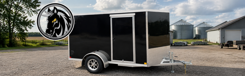 Black enclosed cargo trailer on a gravel lot, with a circular horse-head logo on the front left; farm buildings and silos are in the background.