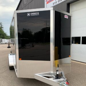 Enclosed black cargo trailer with aluminum trim and diamond plate front, attached to a hitch at a service lot.