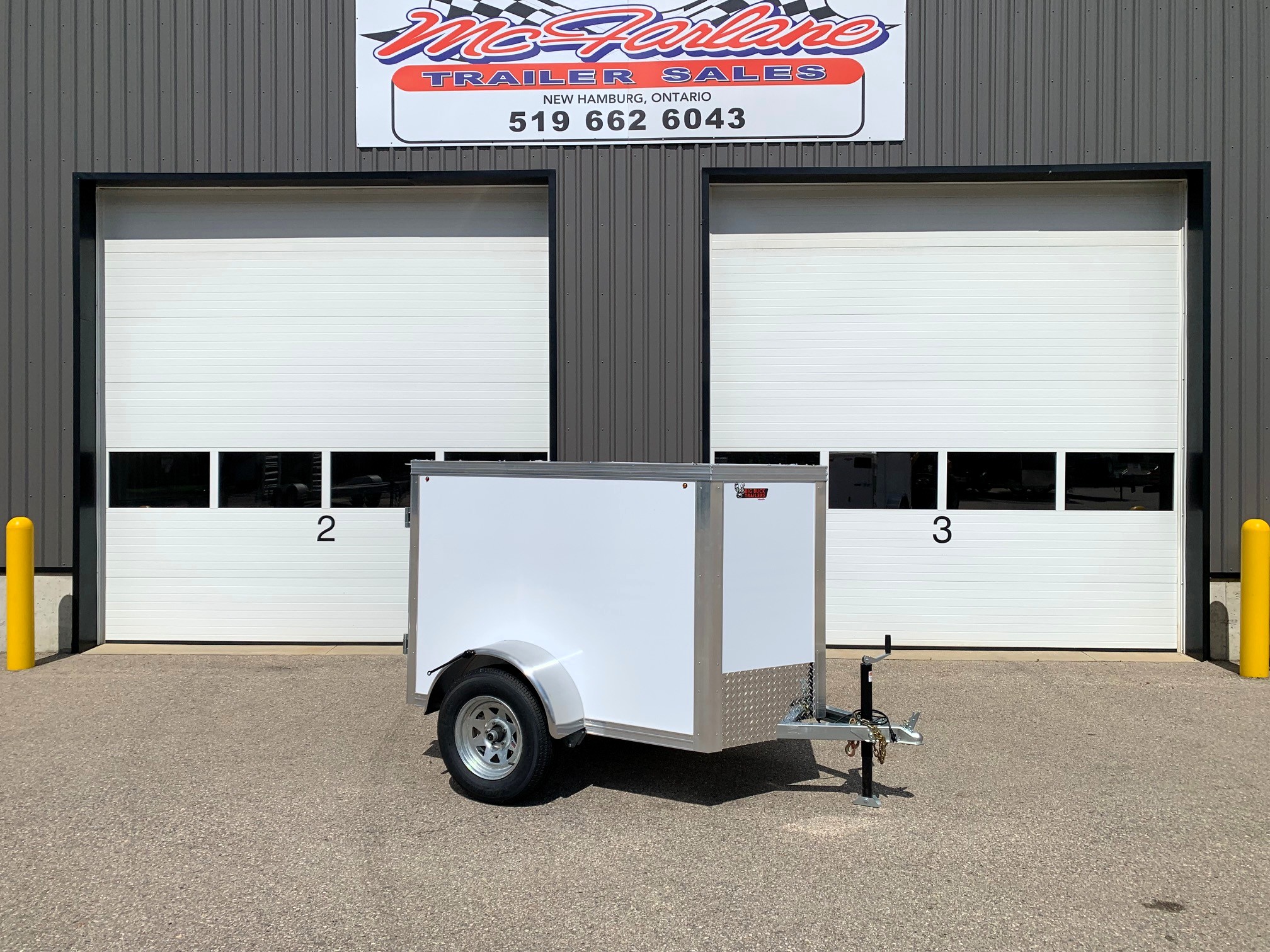 image0 62 - McFarlane Trailer Sales Facade of a gray industrial building with two white roll-up doors; a small silver enclosed trailer parked in front. Above is a sign reading 'McFarlane Trailer Sales, New Hamburg, Ontario, 519-662-6043'.