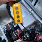 image0 63 - McFarlane Trailer Sales Close-up of a yellow hydraulic control pendant with four round buttons labeled Up/Down Jack and Up/Down T-Box, surrounded by cables in a metal utility compartment.