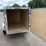 Rear of an empty enclosed trailer with diamond-plate frame and plywood walls, doors open in a parking lot.