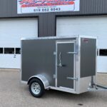 Grey enclosed utility trailer with diamond-plate front, parked in front of a trailer sales building.