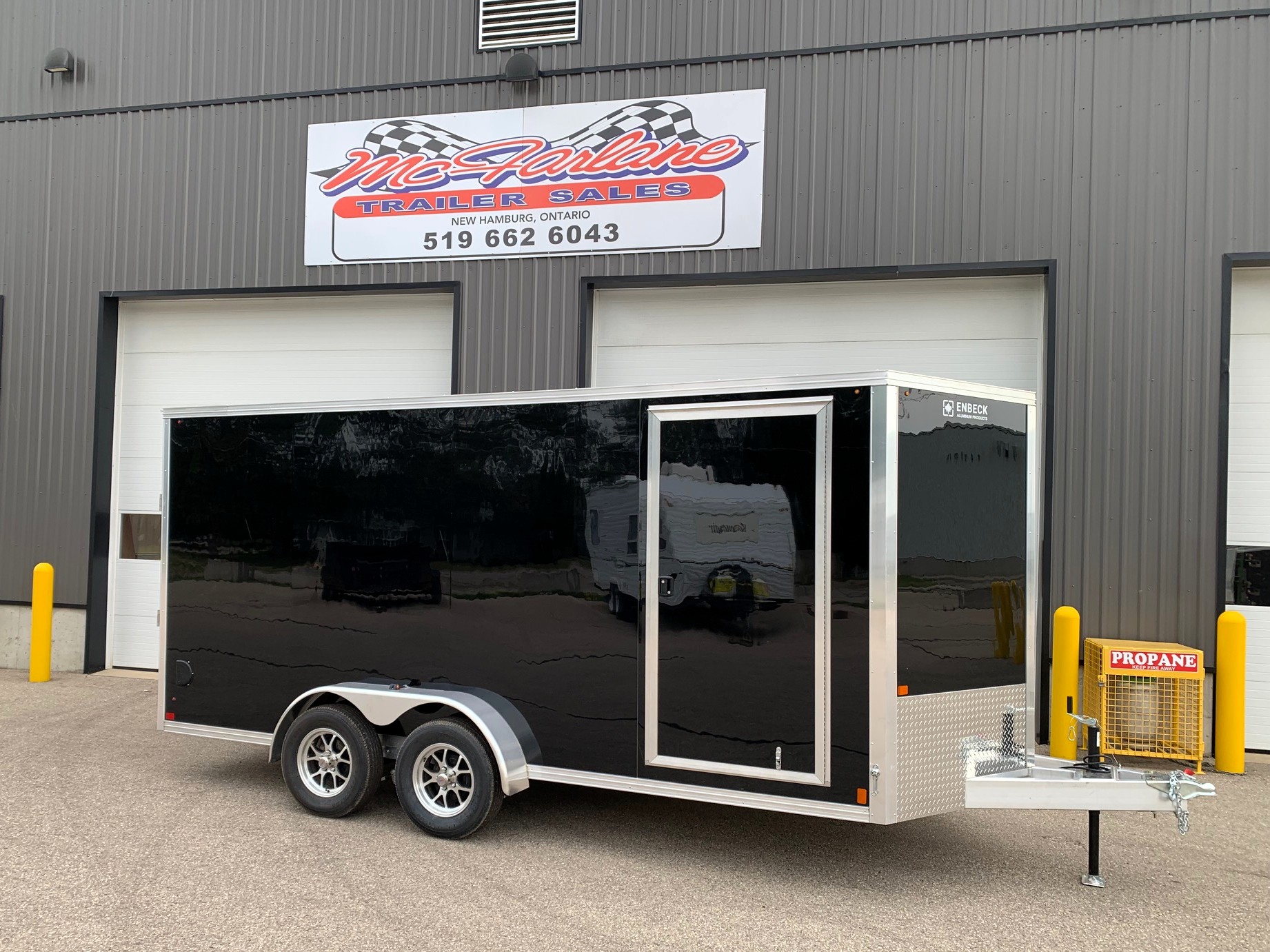 image1 60 - McFarlane Trailer Sales Black enclosed dual-axle cargo trailer with chrome trim and reflective sides, parked in front of a gray industrial building with a 'McAllane Trailer Sales' sign above.