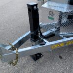 image1 62 - McFarlane Trailer Sales Trailer tongue with a black vertical jack post, safety chains, and a diamond-plate toolbox on a silver trailer labeled DOUGLAS.