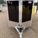 Front view of a black enclosed trailer with aluminum trim and diamond-plate lower front, attached to a hitch with chains on a paved lot.