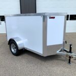 Silver enclosed utility trailer with diamond-plate front, hitch, and 'Big Buck Trailers' logo parked in front of a white building with large doors.