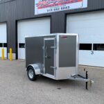 Enclosed gray trailer with diamond-plate front parked in a lot in front of a dark metal building with large white garage doors and a sign above.