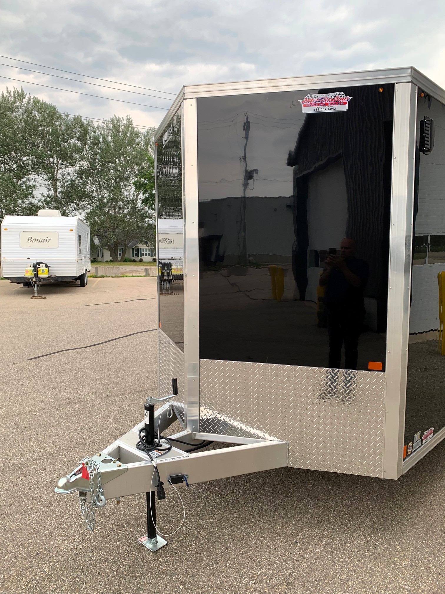 image2 57 - McFarlane Trailer Sales Shiny black enclosed trailer with diamond-plate bottom and hitch in foreground; white Bonair trailer visible in background under a cloudy sky