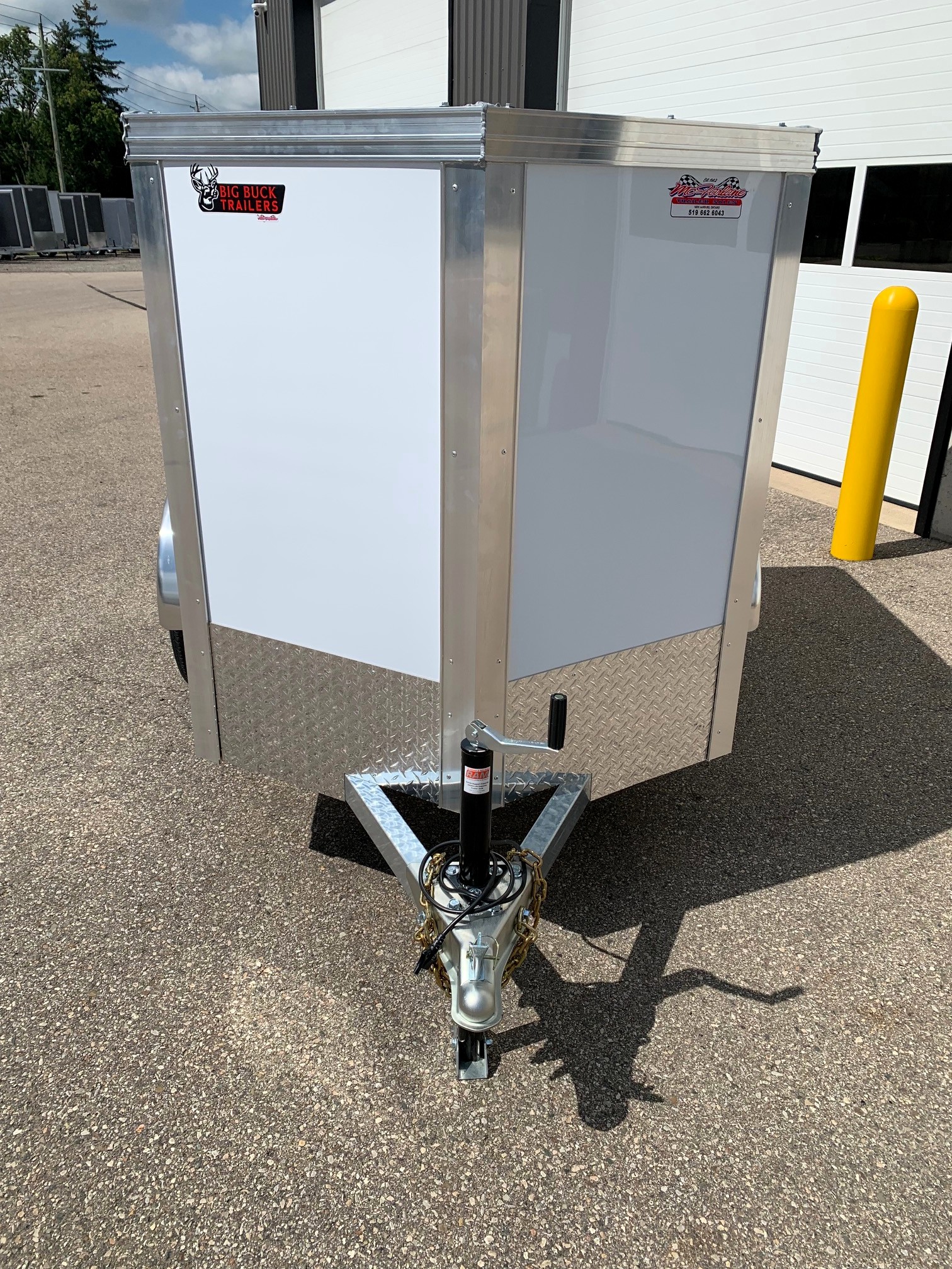 image2 58 - McFarlane Trailer Sales Front view of a silver enclosed utility trailer on a hitch, with diamond-plate at the base and safety chains; outdoors beside a white building.
