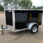 Black enclosed cargo trailer with silver trim and front hitch parked in a lot; safety chains and wheel visible in foreground