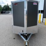 Front view of a gray enclosed cargo trailer with aluminum trim and diamond-plate lower panel, attached to a tow hitch in a parking lot.