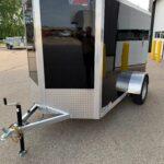 Black enclosed cargo trailer with aluminum trim and diamond-plate front, hitched to a trailer hitch in a parking lot.