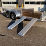Aluminum/open flatbed trailer with wooden deck and diamond-plate ramps extended, parked outside a service bay labeled 2 and 3.