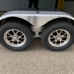 image3 52 - McFarlane Trailer Sales Dual-axle trailer with silver curved wheel fenders and large black tires, viewed at ground level.