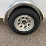 image3 53 - McFarlane Trailer Sales Trailer wheel with a silver rim and black tire under a curved metal fender, attached to a light-colored trailer.