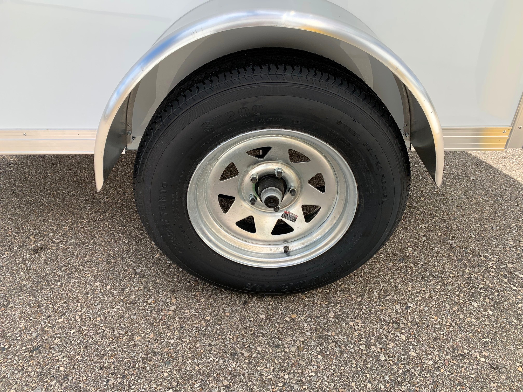 image3 53 - McFarlane Trailer Sales Trailer wheel with a silver rim and black tire under a curved metal fender, attached to a light-colored trailer.
