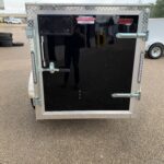 Rear of an enclosed cargo trailer with diamond-plate trim, black door, and metal latches, parked in a lot with tires in the background.