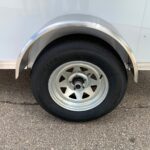 Close-up of a trailer wheel: silver rim, black tire, with a curved metal fender above it on a light-colored trailer.
