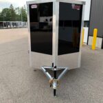 Front view of a black enclosed cargo trailer with aluminum frame and diamond-plate base, hitched in a parking lot outside a building.