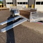 Aluminum utility trailer with two fold-down loading ramps and wooden deck parked in front of a white industrial building with bay doors labeled 1, 2, and 3.