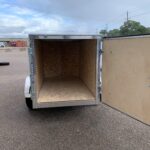Open cargo trailer with diamond-plate exterior and plywood interior in a parking lot, with tires stacked nearby and industrial buildings in the background.