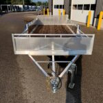 Front view of an empty aluminum utility trailer with diamond-plate sides, attached to a hitch in a loading area near a building with bay doors and yellow bollards.