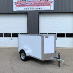 Industrial storefront with two large white roll-up doors; a small silver enclosed trailer is parked in front, hitch tied to a post. Above, a sign reads a trailer sales business name.