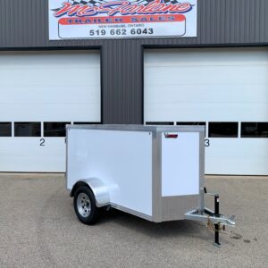 Industrial storefront with two large white roll-up doors; a small silver enclosed trailer is parked in front, hitch tied to a post. Above, a sign reads a trailer sales business name.