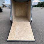 Open cargo trailer with plywood walls and a lowered wooden ramp, ready for loading in a parking lot.
