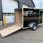 Black enclosed cargo trailer with diamond-plate trim and wooden ramp lowered for loading outside a warehouse.