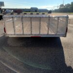 Rear view of a diamond-mesh utility trailer in an industrial parking lot, with stacked tires on the left and warehouses in the background.