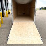 image7 26 - McFarlane Trailer Sales Open box trailer with plywood walls and floor, ramp extended onto a paved loading area in a commercial lot. Yellow bollards and warehouse buildings are visible nearby.