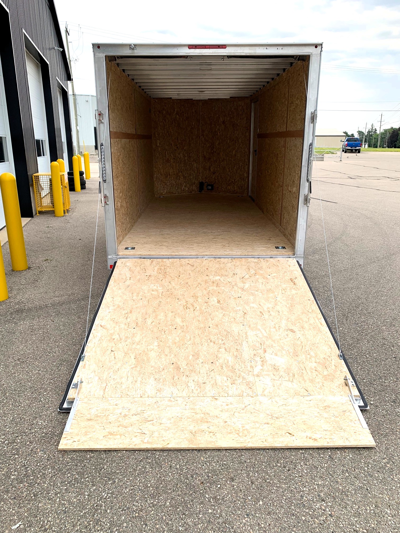 image7 26 - McFarlane Trailer Sales Open box trailer with plywood walls and floor, ramp extended onto a paved loading area in a commercial lot. Yellow bollards and warehouse buildings are visible nearby.