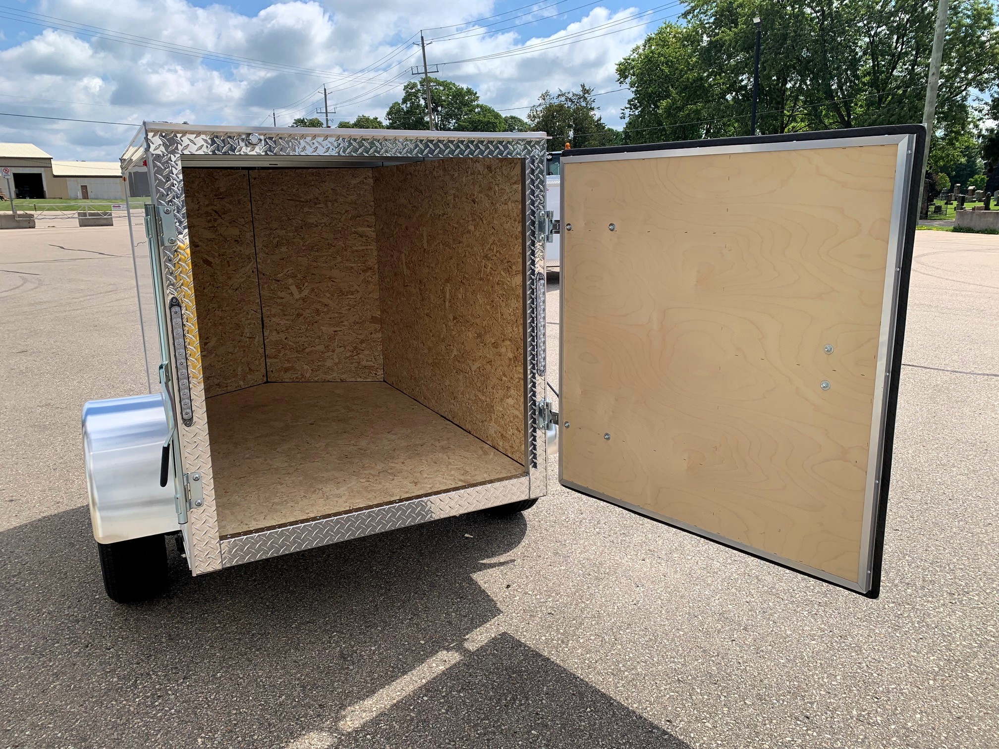 image7 27 - McFarlane Trailer Sales Open cargo trailer with plywood interior and diamond-plate trim along the edges in a parking lot, rear doors swung open.