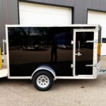 Shiny black enclosed trailer with a silver frame parked in front of a warehouse with white garage doors; foreground shows a wheel and hitch.