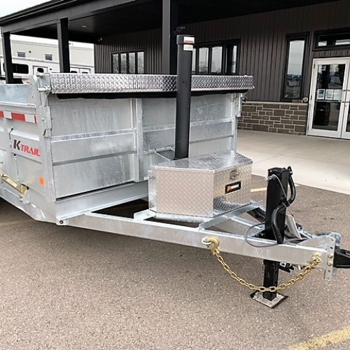 In Content How Many Fall Cleanups Fit in One Load A K-Trail Dump-Trailer Capacity Planner for Landscaping Crews - McFarlane Trailer Sales Silver K-Trail dump trailer with a diamond-plate toolbox and a black hoist/post at the front, parked on a lot in front of a building.