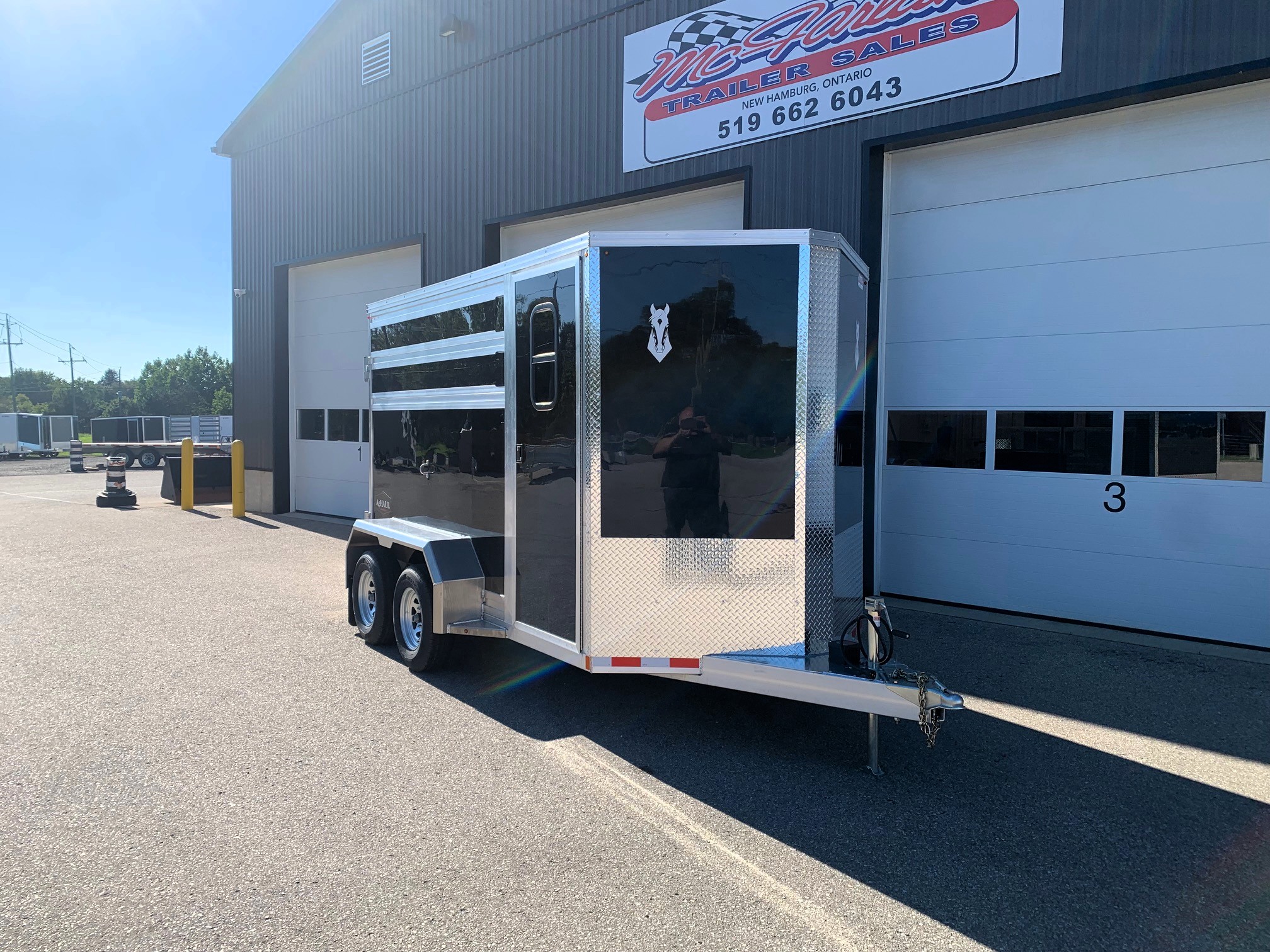 image0 69 - McFarlane Trailer Sales Glossy black enclosed trailer with aluminum trim and diamond-plate accents, parked outside a service building with a large sign.