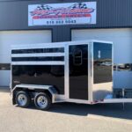 image1 67 - McFarlane Trailer Sales Two-axle black utility trailer with aluminum trim and ramp, parked in front of a gray industrial building with a storefront sign above me.