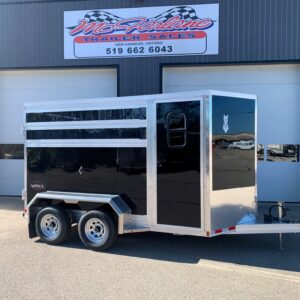 Two-axle black utility trailer with aluminum trim and ramp, parked in front of a gray industrial building with a storefront sign above me.