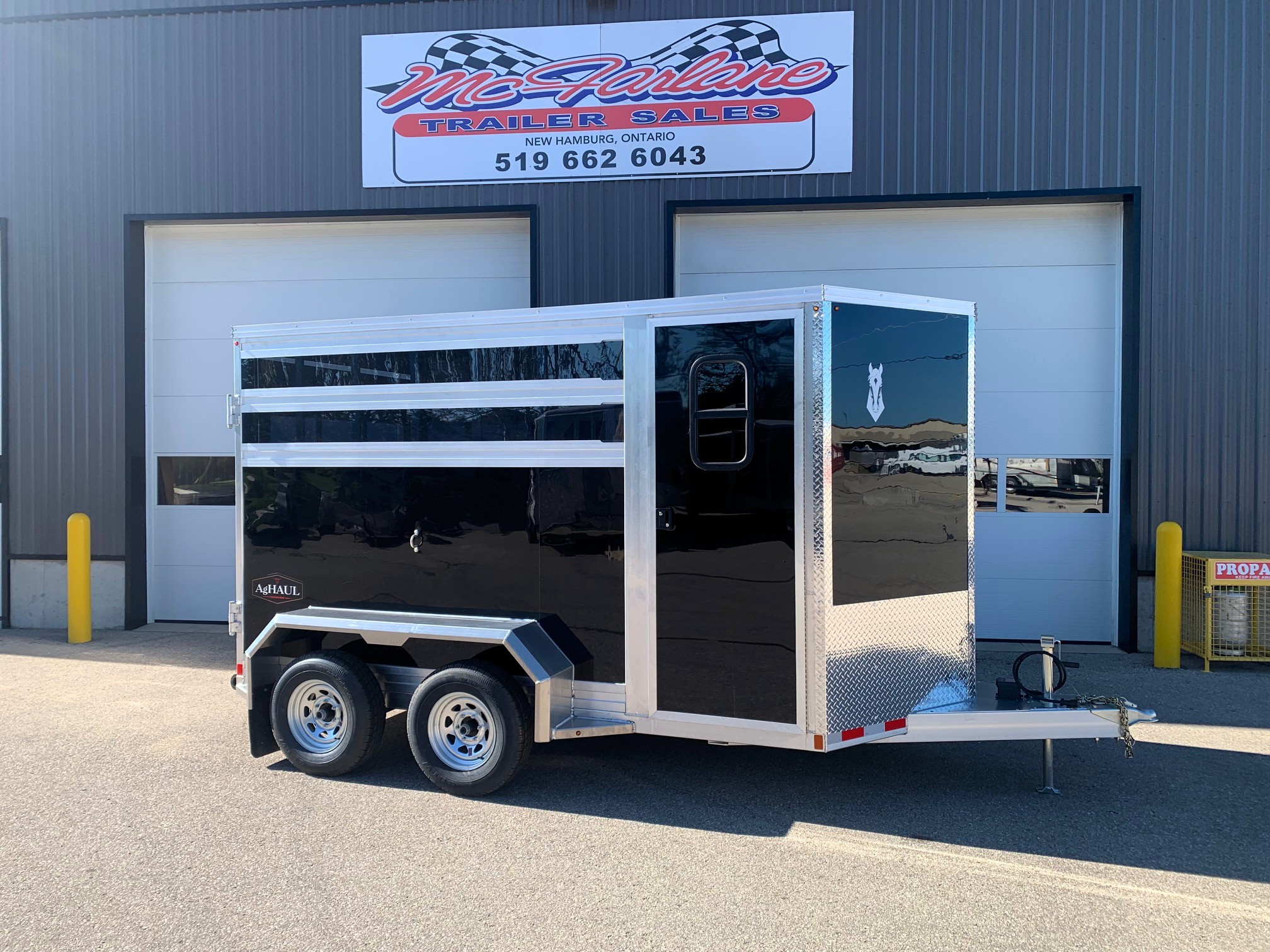 image1 67 - McFarlane Trailer Sales Two-axle black utility trailer with aluminum trim and ramp, parked in front of a gray industrial building with a storefront sign above me.