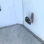 image10 8 - McFarlane Trailer Sales Close-up of a pale blue metal door showing a silver latch plate and mounting bracket on the right, with a dark curved handle on the left side.