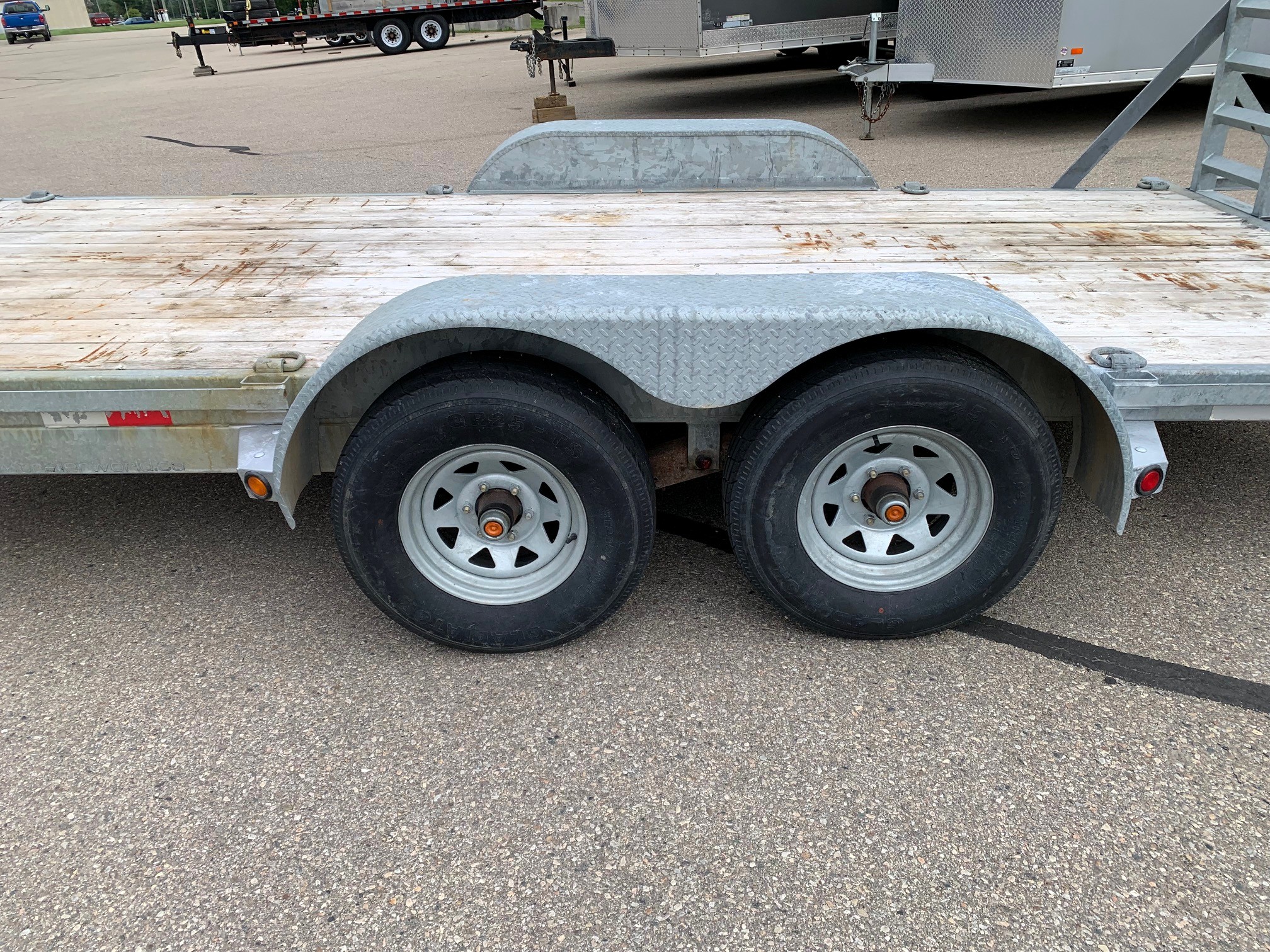 image2 65 - McFarlane Trailer Sales Dual-axle trailer with a weathered wooden deck and curved steel fenders, parked on asphalt with other trailers in the background.