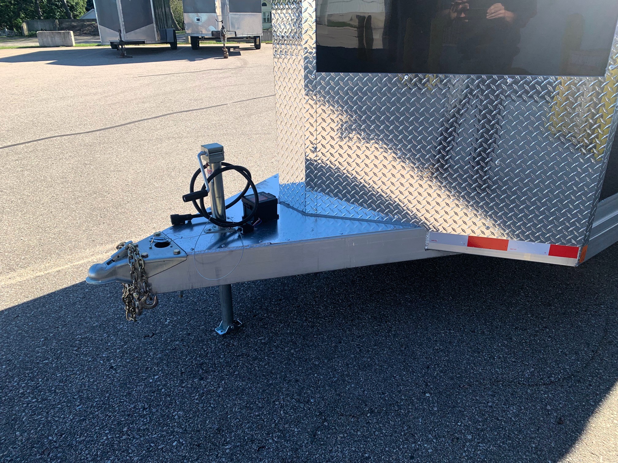 image3 59 - McFarlane Trailer Sales Close-up of a trailer tongue with a hitch, electrical plug and chains on a diamond-plate trailer in a parking lot.