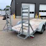 image3 60 - McFarlane Trailer Sales Metal utility trailer with tall metal rack frames on a wooden deck in a paved lot near a gray industrial building with large doors.