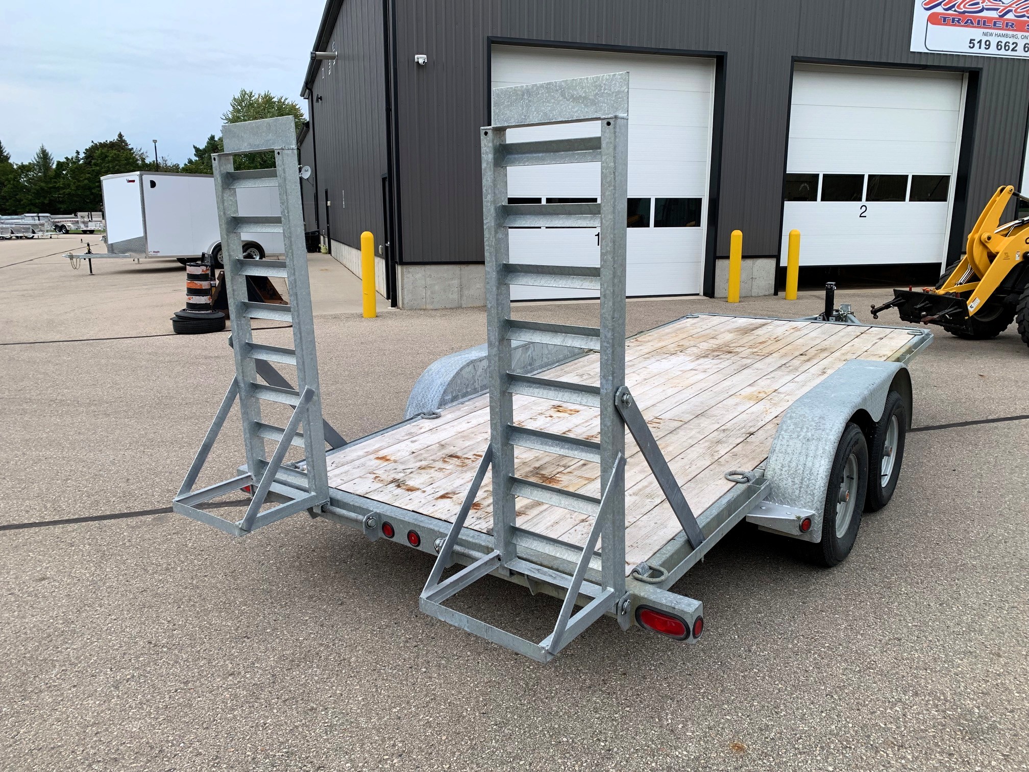 image3 60 - McFarlane Trailer Sales Metal utility trailer with tall metal rack frames on a wooden deck in a paved lot near a gray industrial building with large doors.