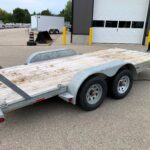 image4 58 - McFarlane Trailer Sales Flatbed utility trailer with wooden deck and dual axles parked in a lot near a warehouse, with yellow bollards and other trailers in the background.
