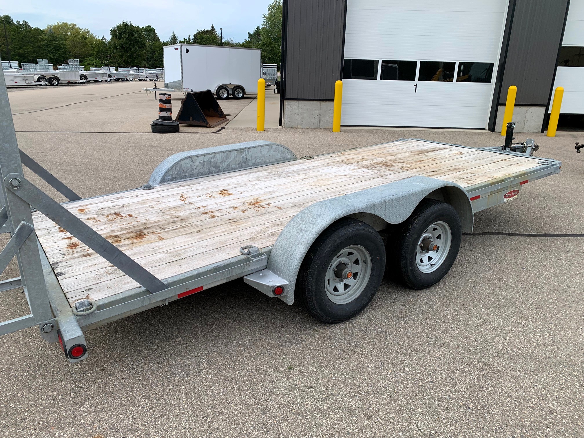 image4 58 - McFarlane Trailer Sales Flatbed utility trailer with wooden deck and dual axles parked in a lot near a warehouse, with yellow bollards and other trailers in the background.