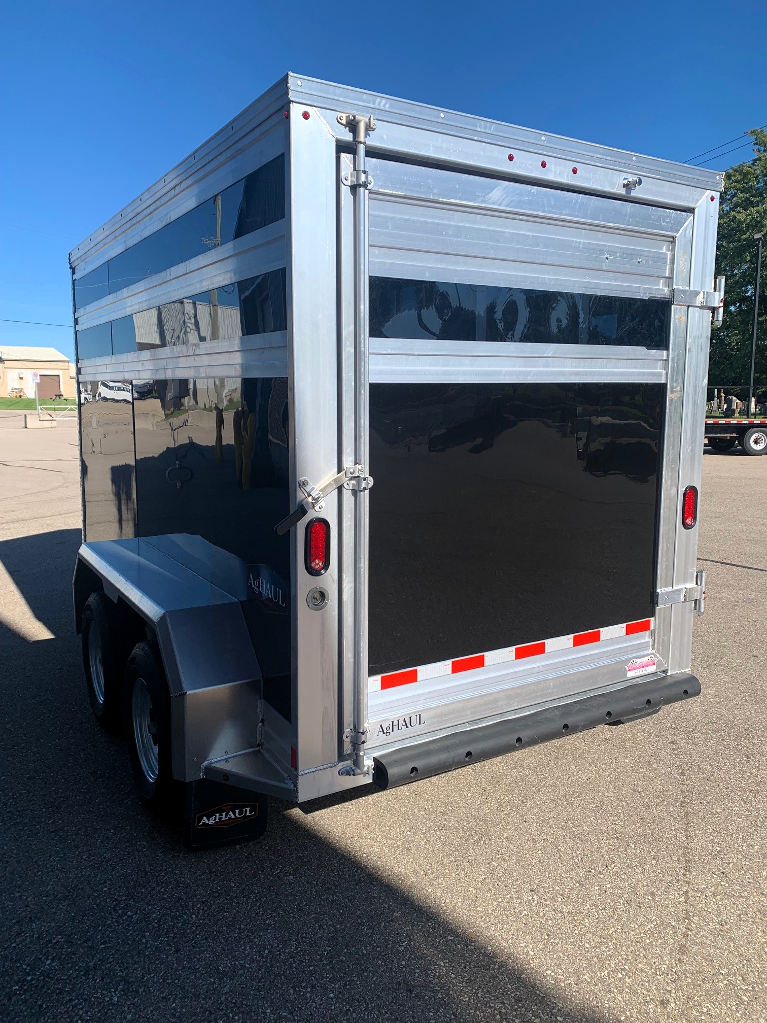 image5 50 - McFarlane Trailer Sales Rear of a silver aluminum cargo trailer with a black back panel, red tail lights, and red-white reflective stripes along the lower edge.